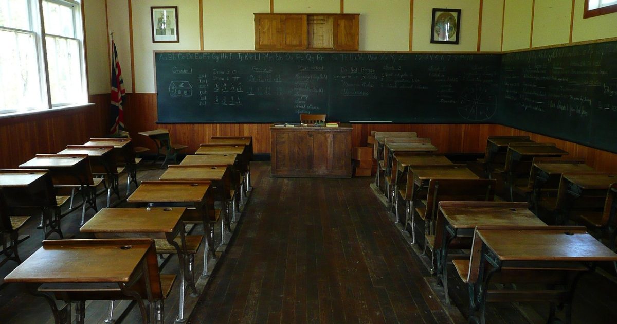 复古暖色调的教室场景中，教师手持平板演示极域电子教室 App 的学生端互动界面，木桌上散放着粉笔与笔记本，阳光斜照进窗格